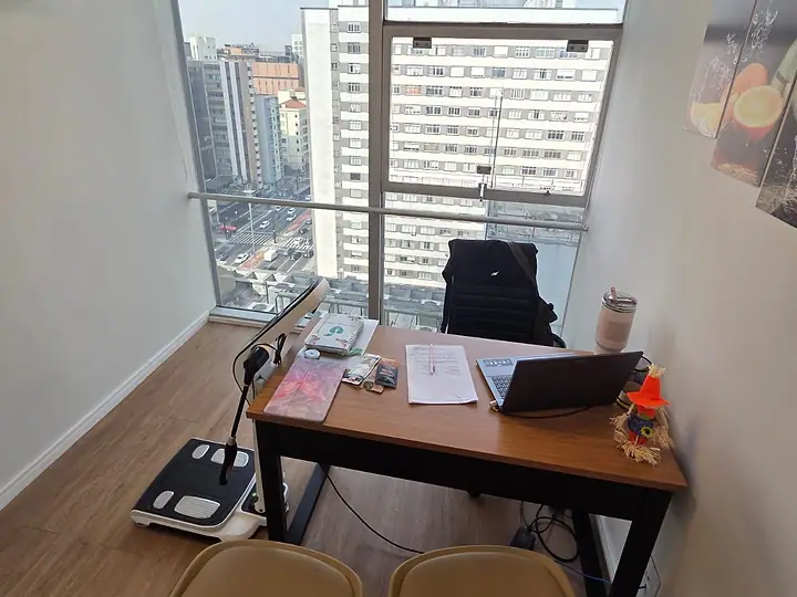 Sala do consultório da Av. Paulista, com mesa, duas poltronas e janela para os prédios da avenida