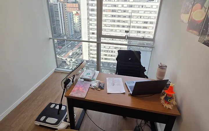 Sala do consultório da Av. Paulista, com mesa, duas poltronas e janela para os prédios da avenida