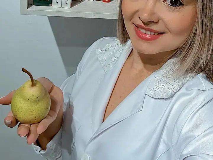 Natacha Ludkiewicz sorrindo, de jaleco, com uma pera na mão, diante de uma estante de livros
