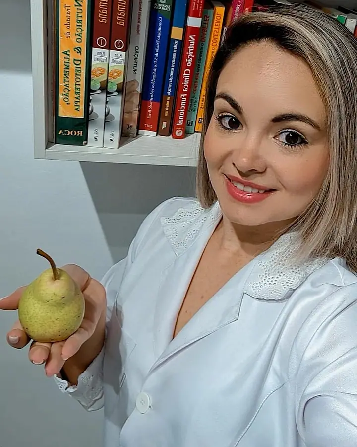 Natacha Ludkiewicz sorrindo, de jaleco, com uma pera na mão, diante de uma estante de livros